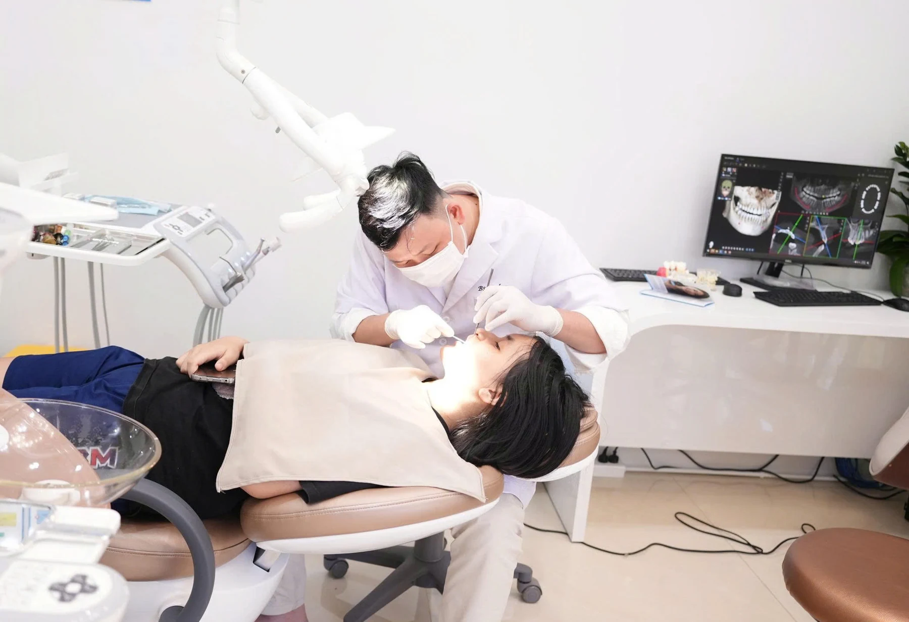 Nhân viên y tế điều trị cho người bệnh tại ghế nha khoa, bên cạnh màn hình quét 3D